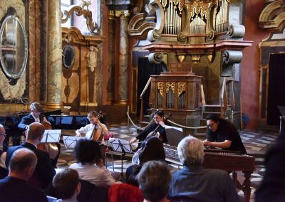 Adventní koncert ZUŠ 7.12.2019 - Klementinum. Pohled na instrumentální soubor, děvče hrající na cimbál, paní učitelka D. Pražáková hrající na violoncello a dva chlapci hrající na violoncello.