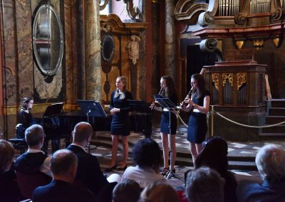 Adventní koncert ZUŠ 7.12.2019 - Klementinum. Pohled na zpívající děvče, děvče hrající na klavír a dvě děvčata hrající na flétnu.