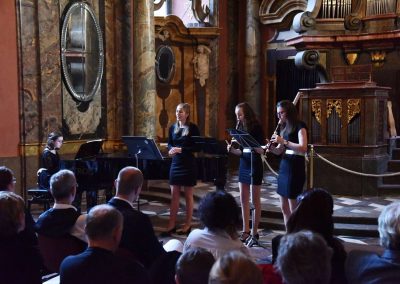 Adventní koncert ZUŠ 7.12.2019 - Klementinum. Pohled na zpívající děvče, děvče hrající na klavír a dvě děvčata hrající na flétnu.