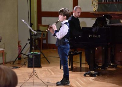 Refektář profesního domu MFF UK, dechový koncert 6.2.2020. Žák hrajíci na zbocovu flétnu - chlapec, v pozadí učitel hrající na klavír.