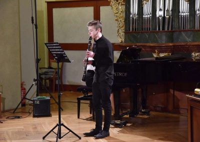Refektář profesního domu MFF UK, dechový koncert 6.2.2020. Žák hrajíci na klarinet - chlapec, v pozadí učitelka hrající na klavír.