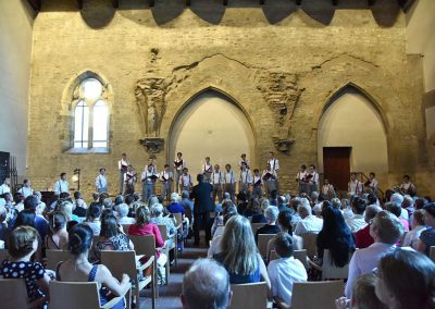 Závěrečný koncert Pueri gaudentes 24.6.2019 - Anežský klášter. Pohled na publikum, pana sbormistra L. Sládka a zpívající mužský sbor.