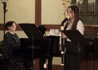 Dechový koncert, Profesní dům MFF, žačka hrající na zobcovou flétnu a paní učitelka doprovázející na klavír.