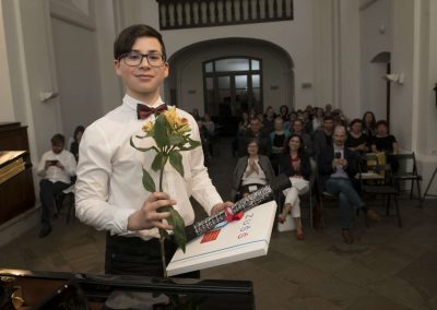 I. absolventský koncert, Atrium na Žižkově, 10.5.2023, absolvent s kytkou a vysvědčením a za ním publikum