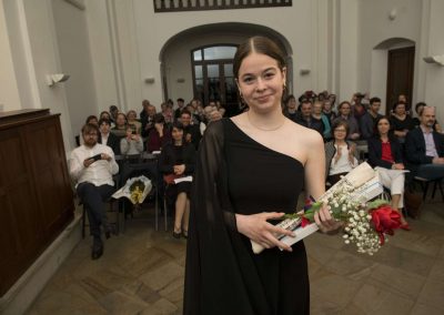 I. absolventský koncert, Atrium na Žižkově, 10.5.2023, absolventka s kytkou a vysvědčením a za ní publikum