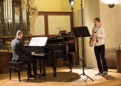 Dechový koncert, Profesní dům MFF, 27.2.2023, žák hrající na saxofon a pan učitel